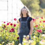 Woman in denim overalls standing in a field of flowers with a greenhouse behind her.