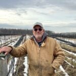 Man dressed in a beige coat and ball cap standing in a farm field in winter