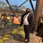 Man standing in front of window, standing beside a poster of a magazine cover with his own face on it