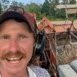 Picture of a smiling man with headphones and a hat. Behind him farm machinery is harvesting crops.