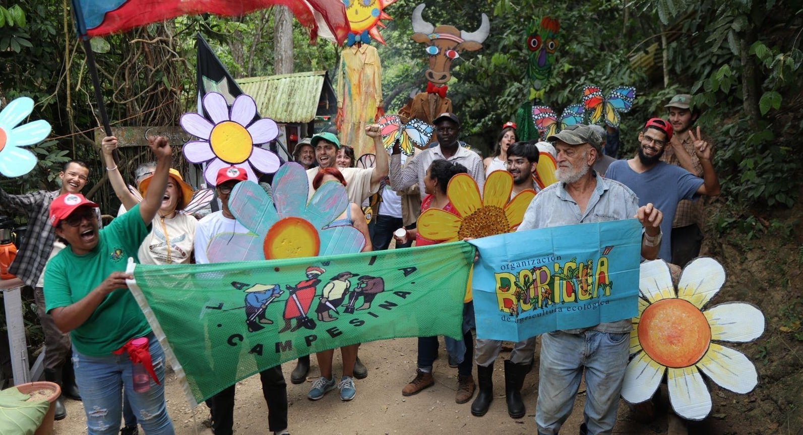 There is a parade of people on a path through the jungle. They are holding large cardboard flowers and flags.